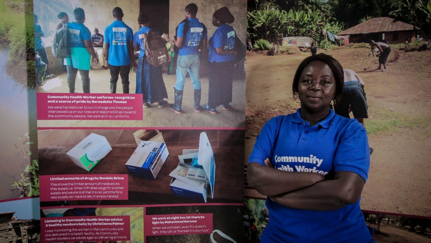 Bernadette Thomas a Community Health Worker from Bonthe District of Sierra Leone poses next to a photo exhibition about the challenges she and her colleagues face in the course of their work.