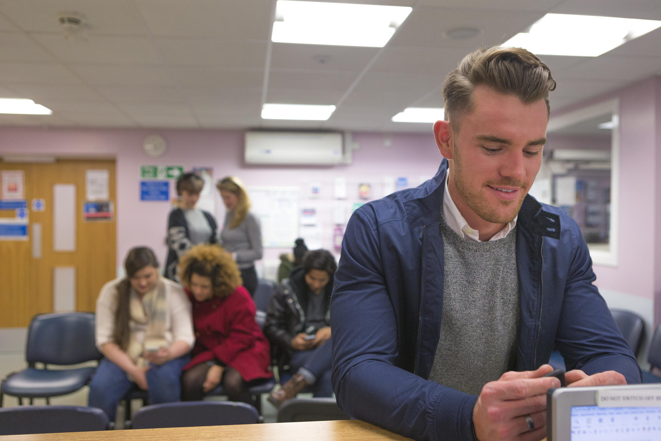 A horizontal image of a man in a doctor's clinic smiling at the receptionist booking him in for a medical check. He looks very cheerful and positive.